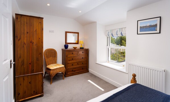 A bedroom with a wooden wardrobe, a wicker chair, a dresser, a window with a view, and a framed picture on the wall. - Fern Cottage (Ambleside)