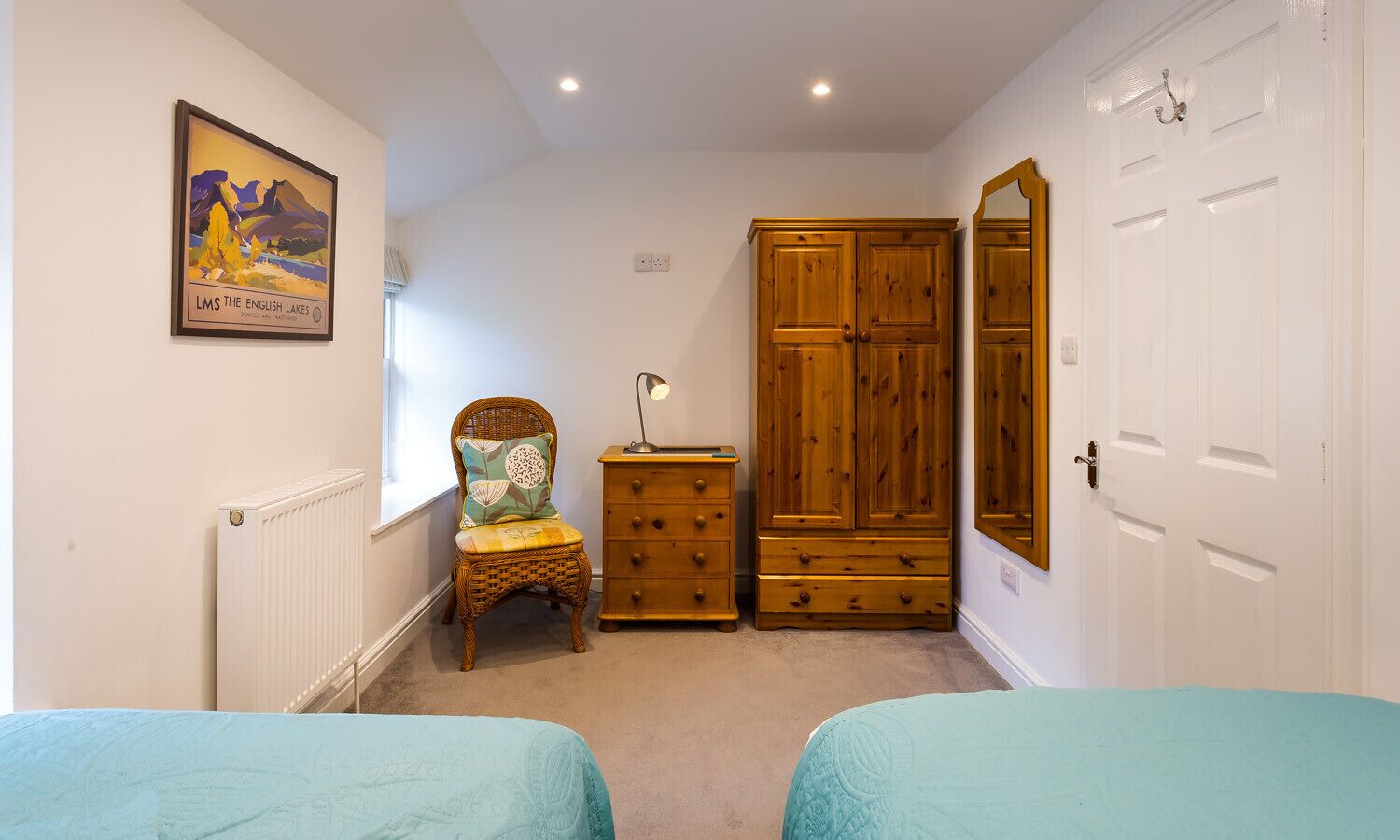 A bedroom with two beds, a wooden wardrobe, a chair with a cushion, a radiator, and a framed picture on the wall.