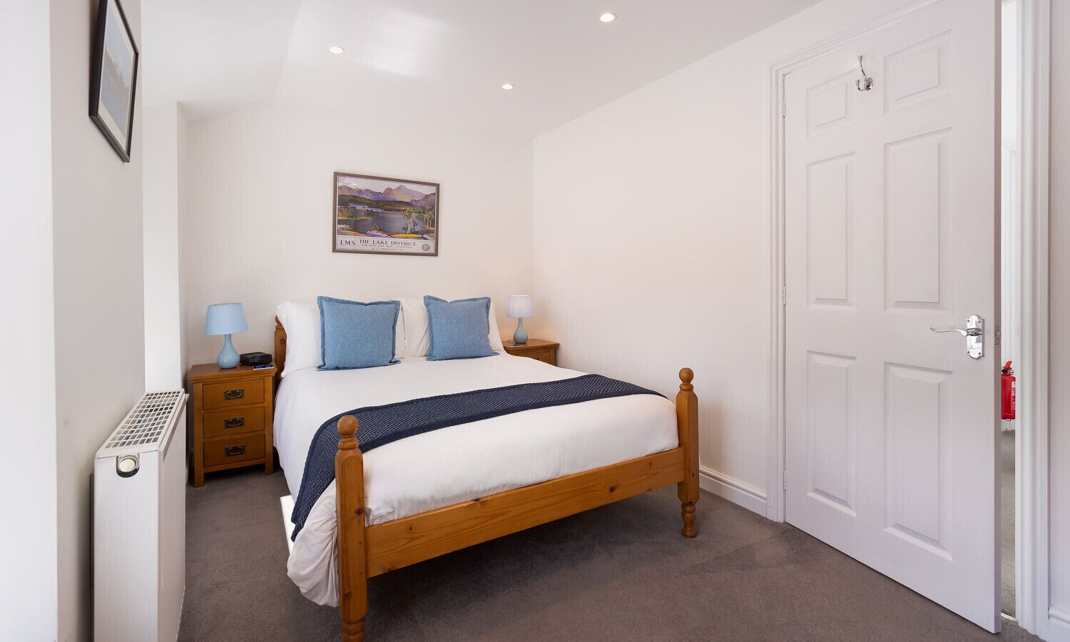 A bedroom with a wooden bed, a nightstand, a lamp, a framed picture on the wall, and a white door.