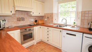 A kitchen with white cabinets, a wooden countertop, a built-in oven, a dishwasher, and a window with a view of greenery. - Millerbeck (Ambleside)