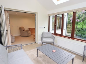 A room with a sofa, a grey armchair, a small table, and a view of a garden through large windows.