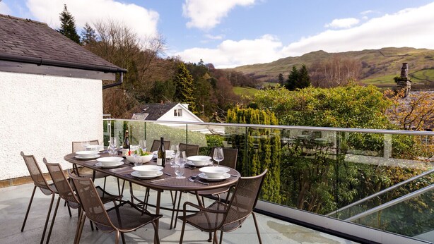 Comedor con mesa preparada para seis personas, sillas exteriores y barandilla de vidrio con vista a un paisaje pintoresco.