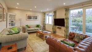 Una sala de estar con un sofá color beige, un sofá de cuero marrón, una mesa de centro de madera y una estantería. Hay un televisor montado en la pared y una ventana con cortinas.