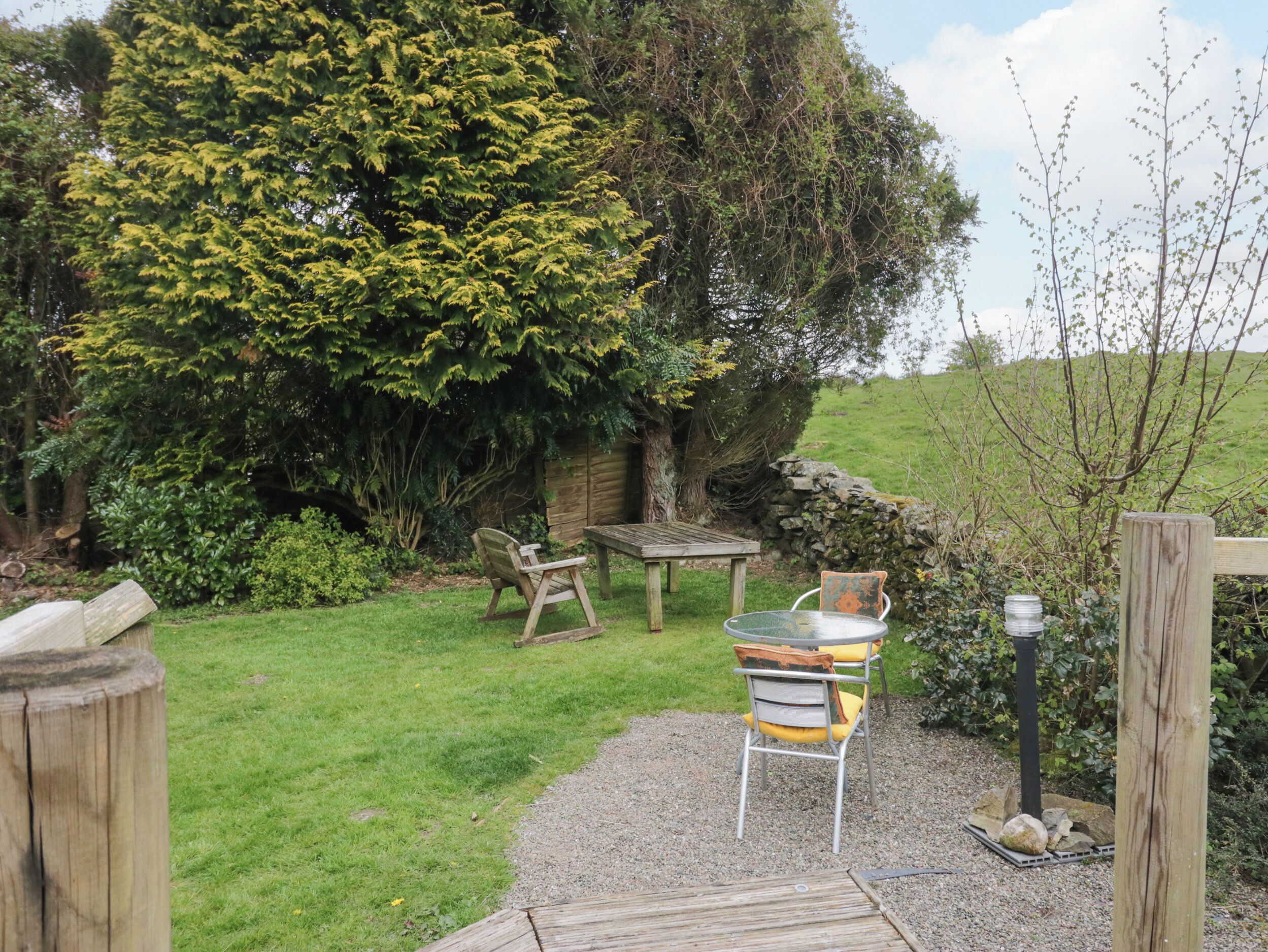 A rustic outdoor seating area with a wooden table and chairs, surrounded by lush greenery and a stone wall.