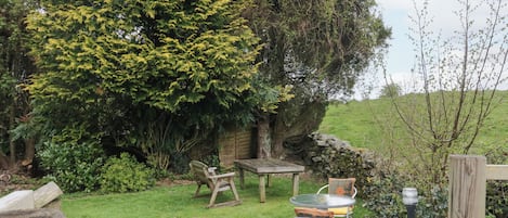 A rustic outdoor seating area with a wooden table and chairs, surrounded by lush greenery and a stone wall.