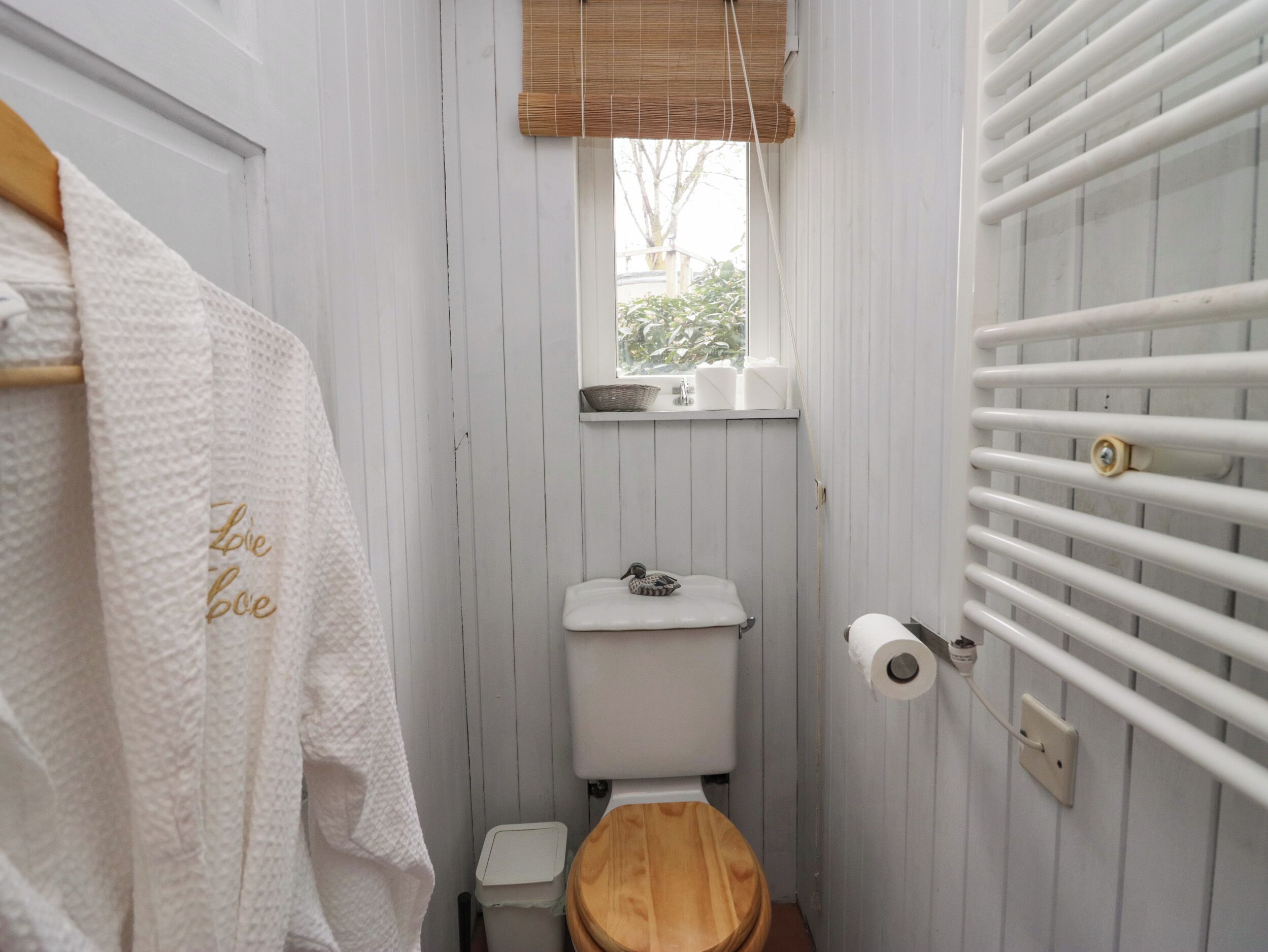 A small bathroom with a wooden toilet, a white towel rack with a towel, a window with a view of greenery, and a white radiator.