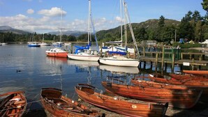 A marina with various boats docked, including sailboats and rowboats, surrounded by a calm lake and hills in the background.