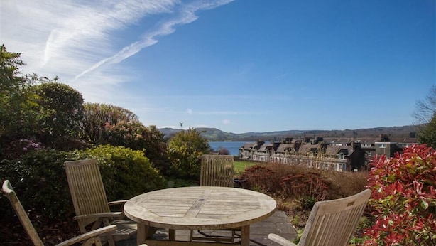 A round wooden table with four chairs in a garden setting, overlooking a lake and residential buildings.