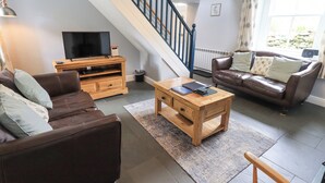Un salon avec un canapé en cuir marron, une table basse en bois, un téléviseur posé sur un meuble en bois, un escalier avec des rampes noires et une fenêtre avec des rideaux.