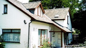 A white house with a sloped roof, a front porch, and a small garden with flowers. - Yew Tree Cottage (Ambleside)