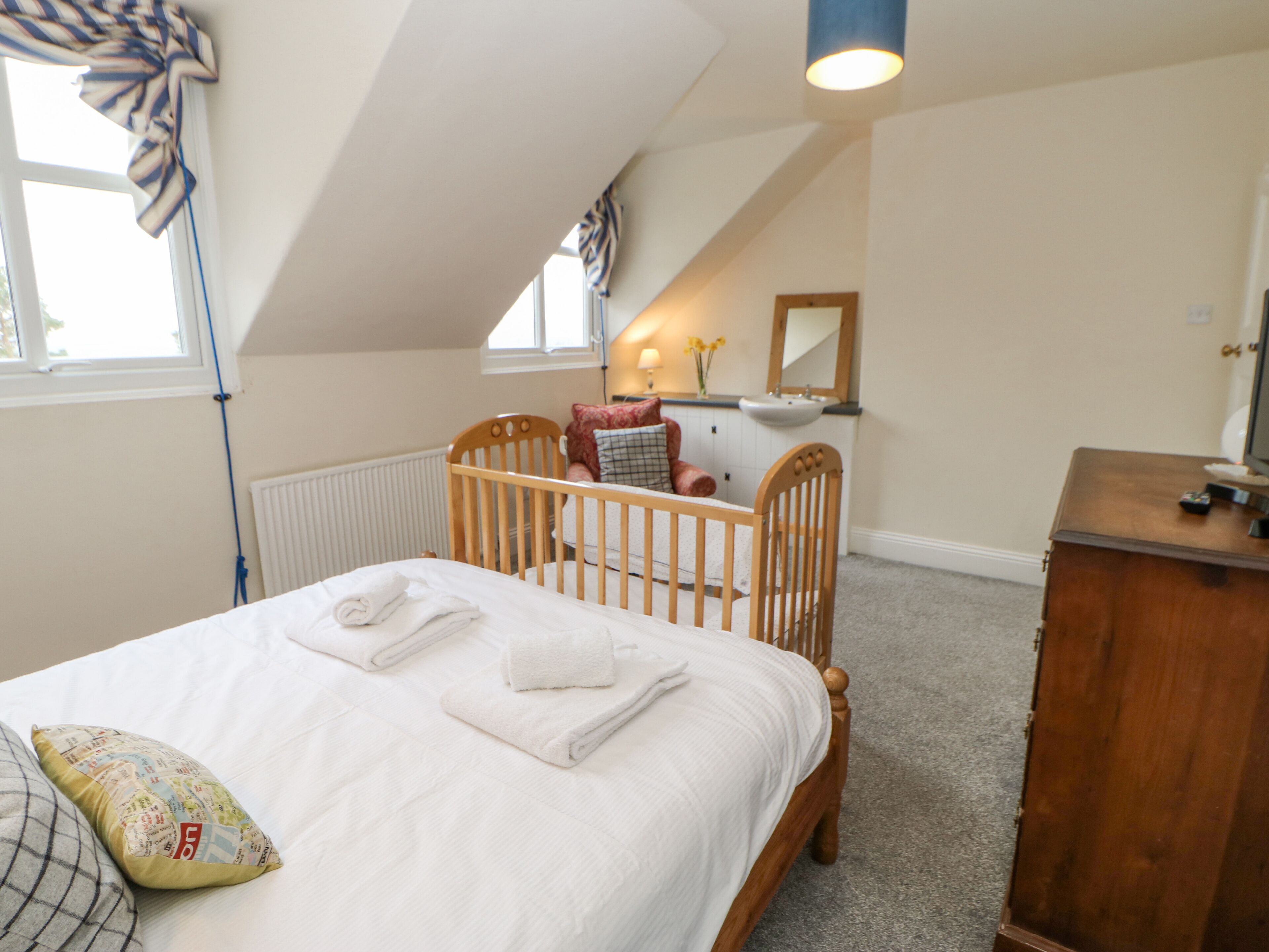A neatly made bed with white linens and towels, a wooden crib, a dresser, and a window with curtains.