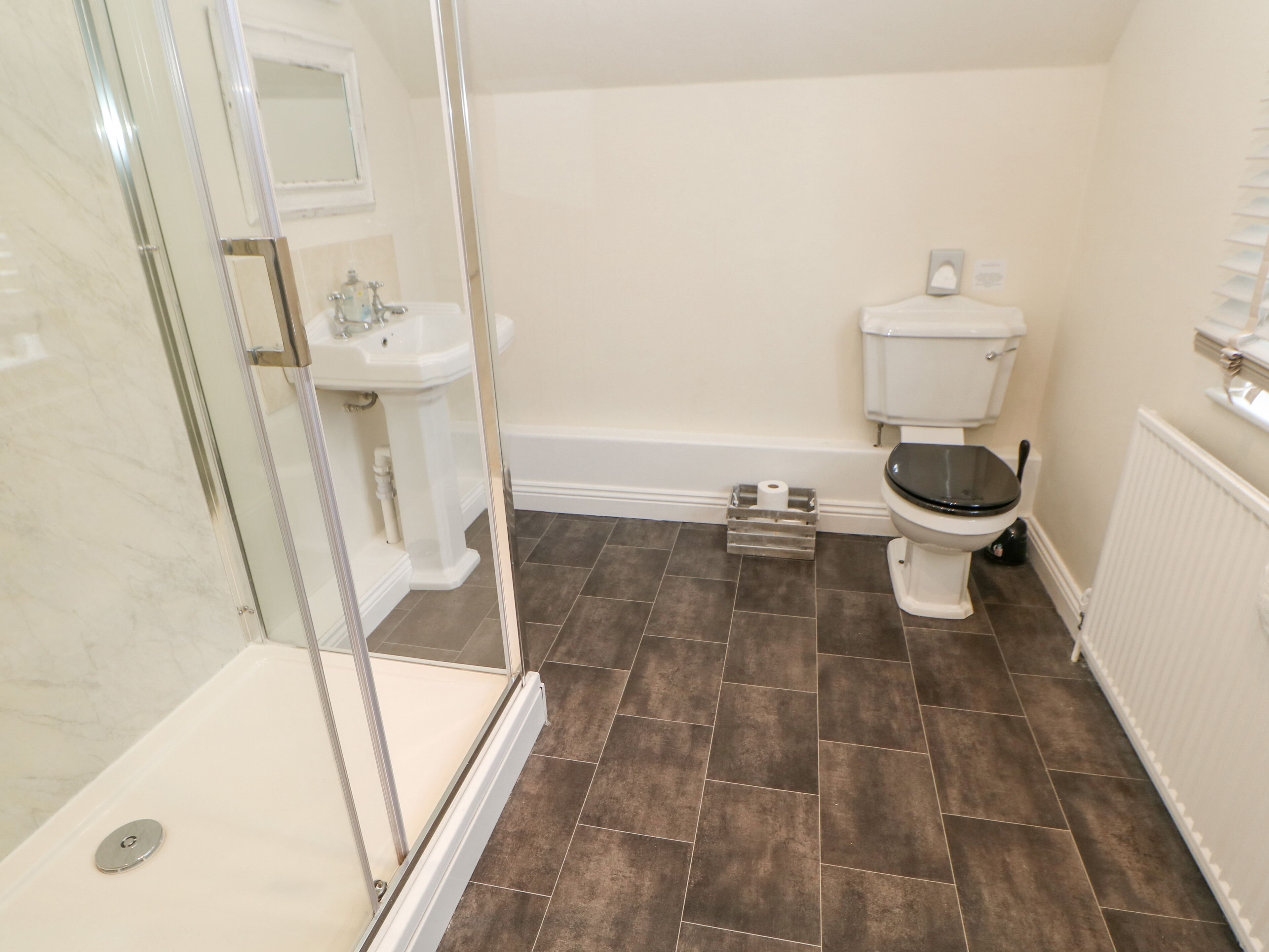 Une salle de bain dotée d’une douche à parois de verre, d’un lavabo sur colonne blanc, de toilettes et d’un carrelage brun foncé.