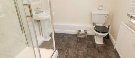 Une salle de bain dotée d’une douche à parois de verre, d’un lavabo sur colonne blanc, de toilettes et d’un carrelage brun foncé.