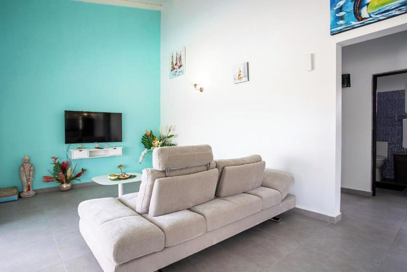 Un salon avec un canapé beige, un téléviseur à écran plat fixé au mur, une petite table d’appoint blanche ornée d’une plante et une statue décorative.