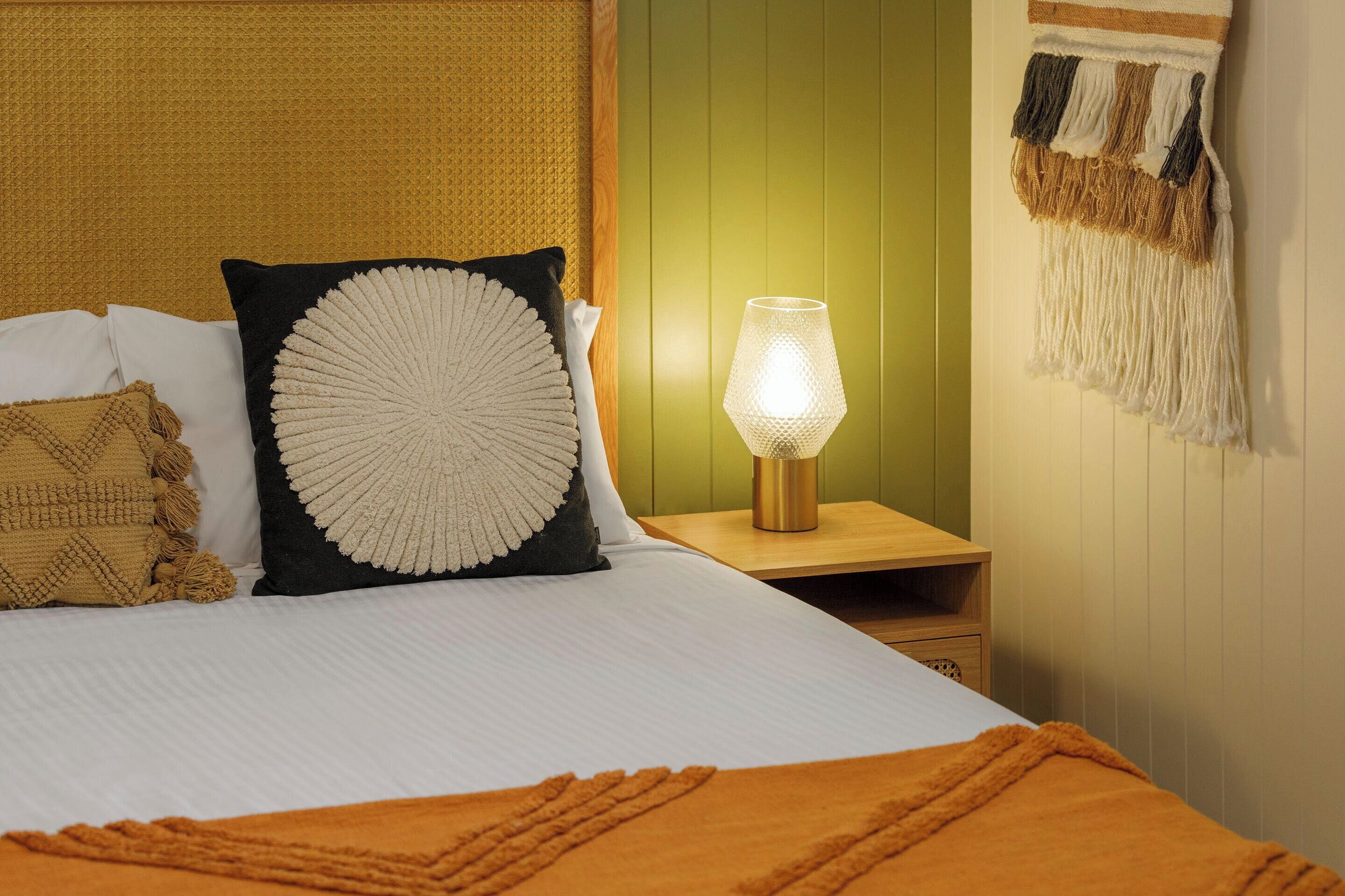 A neatly made bed with a white and orange blanket, a lamp on a bedside table, and a wall with a hanging tapestry.