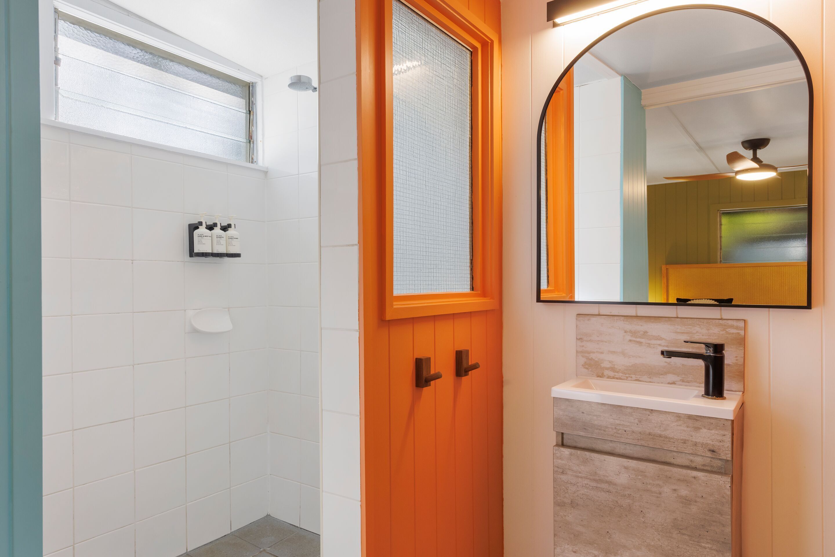 A bathroom with a white tiled wall, a wooden door with two handles, a sink with a black faucet, and a mirror with an arch frame.