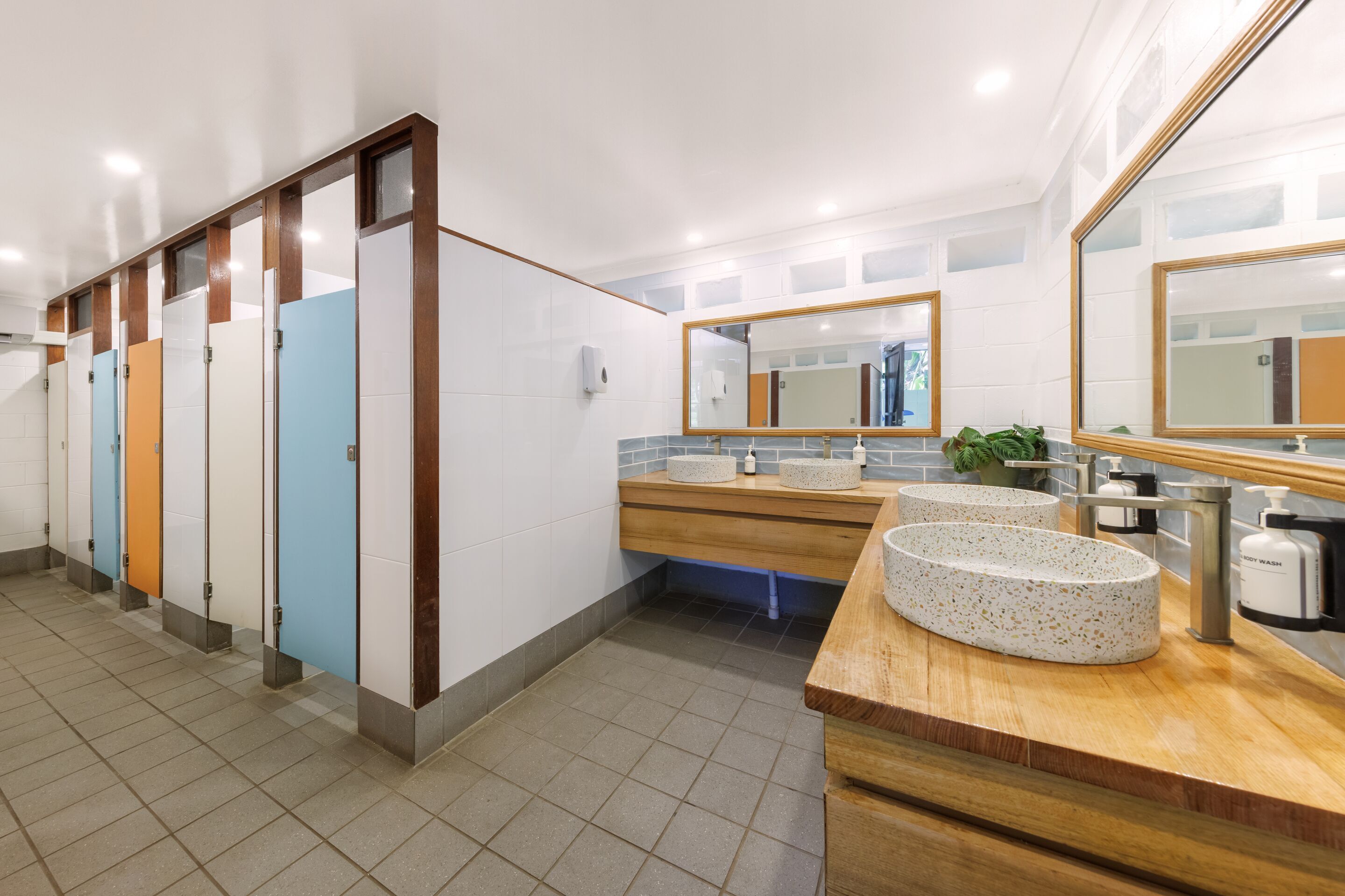 A modern restroom with multiple stalls, a wooden vanity with two sinks, and a large mirror.