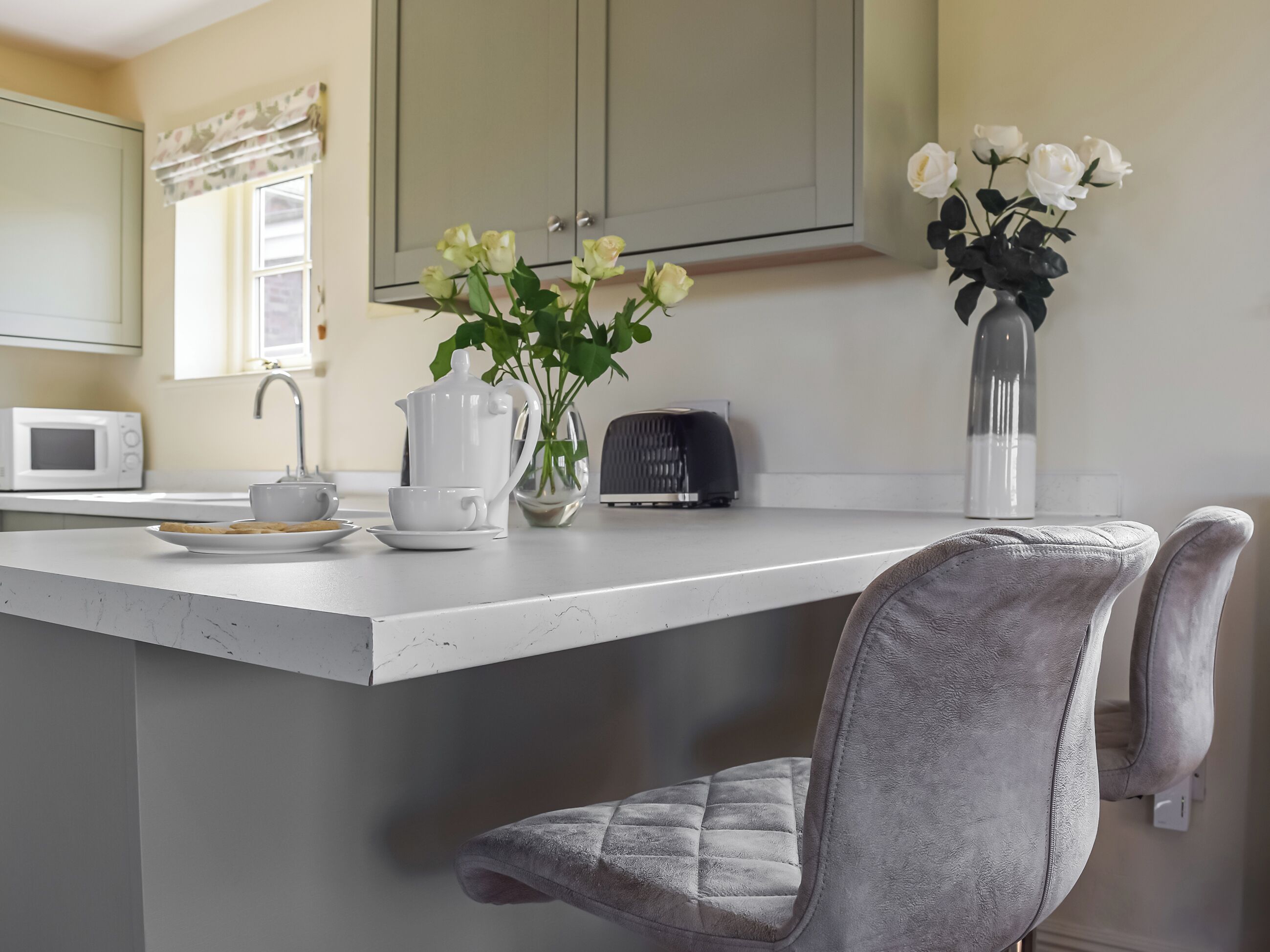 A kitchen with a white countertop, a microwave, a kettle, a vase with flowers, and a chair.