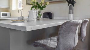 Una cucina con un piano di lavoro bianco, un forno a microonde, un bollitore, un vaso con fiori e una sedia.