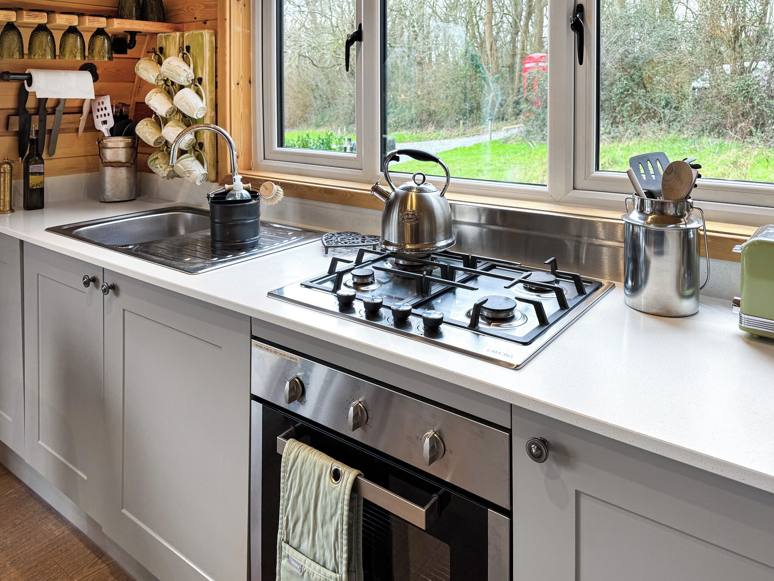 Une cuisine moderne équipée d’une cuisinière à gaz, d’un évier et d’un four, de rayonnages en bois où sont rangés les ustensiles de cuisine, et d’une fenêtre donnant sur un paysage verdoyant.