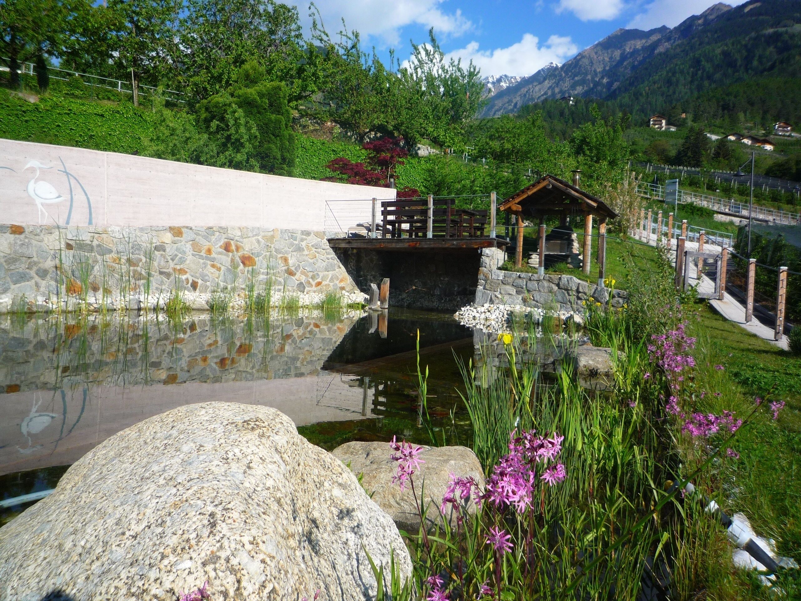 Eine friedliche Seelandschaft mit einem hölzernen Pavillon, einer Steinmauer und freiem Blick auf die umliegenden Berge.
