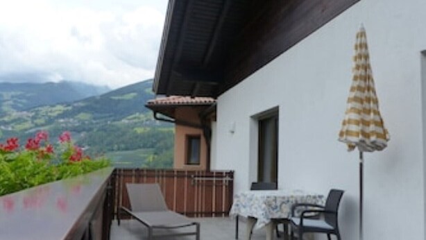Un balcon avec une table et des chaises, une vue sur des collines verdoyantes et un parasol rayé.
