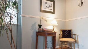 Une pièce d’angle avec un plancher en bois, une chaise avec un coussin, une table avec une lampe, un tableau encadré au mur et une plante en pot.