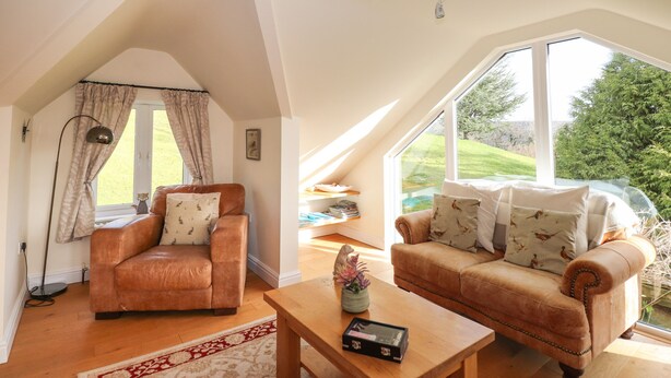 Um quarto de loft aconchegante com teto inclinado, uma janela grande com vista para o exterior, uma poltrona de couro marrom, uma mesa de centro de madeira e um sofá bege.