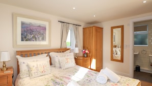 Quarto com cama de madeira, roupa de cama floral, mesa de cabeceira com abajur, guarda-roupa de madeira, janela com cortinas e banheiro visível por uma porta aberta.