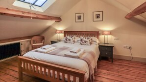 Un dormitorio acogedor en el ático, con techo inclinado, vigas de madera, tragaluces, una cama con ropa de cama floral, dos lámparas de noche y una silla.