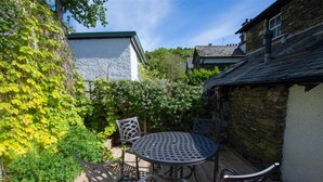 A patio with a round metal table and four chairs, surrounded by greenery and stone walls. - Greystones (Windermere)
