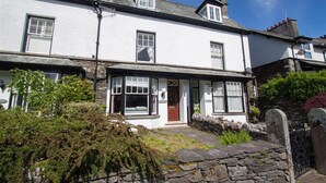 A white two-story house with a stone wall entrance, a front porch, and a small garden area. - Greystones (Windermere)
