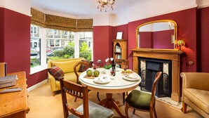 A dining room with a round table, chairs, a fireplace, and a view of the outside. - Greystones (Windermere)