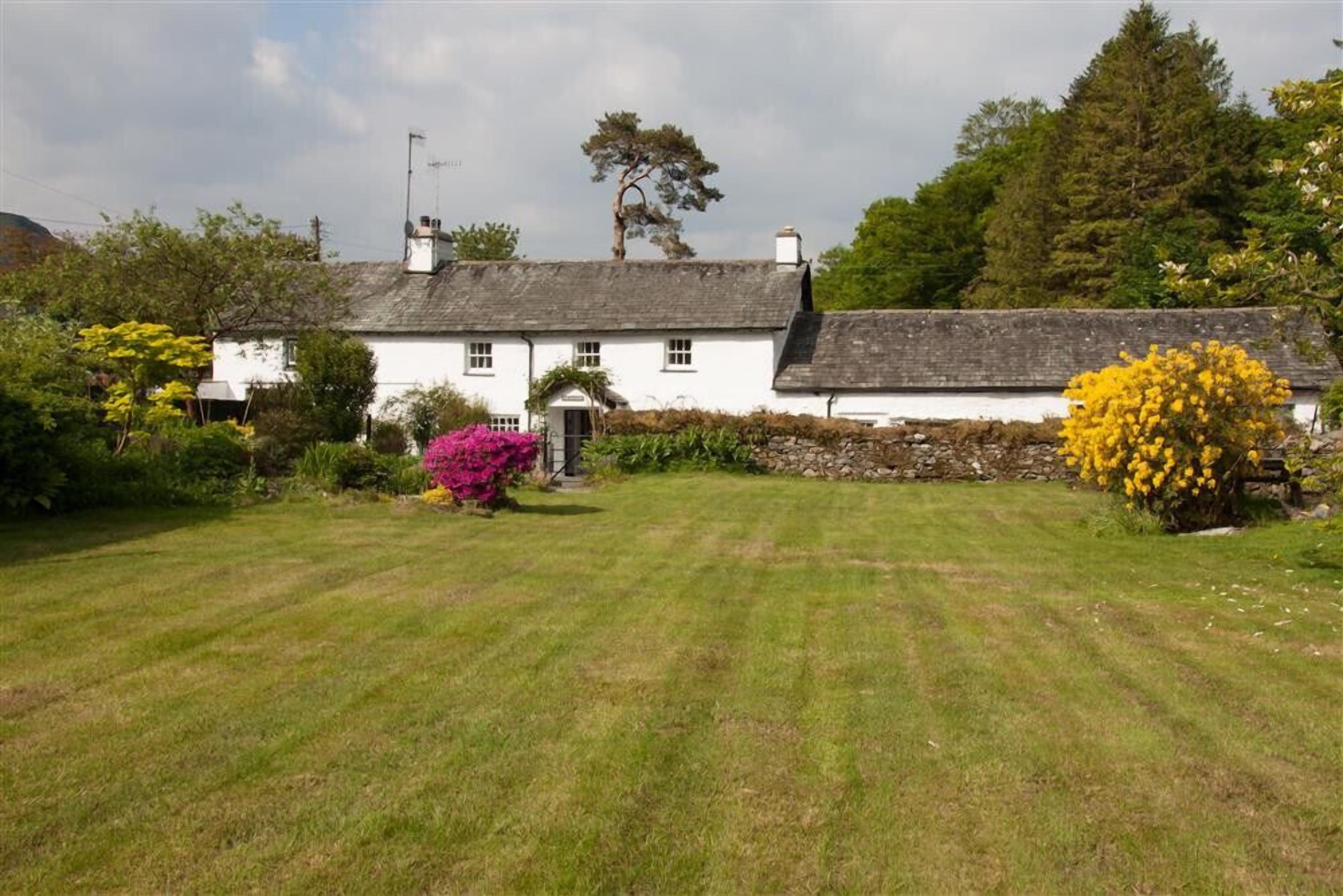 A white house with a grey roof, surrounded by a well-maintained lawn and flowering plants.