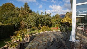 Un patio de piedra con diversas plantas y flores, un seto bien cuidado y una montaña al fondo.