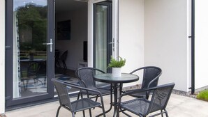 Une terrasse dotée d’une porte vitrée donnant sur une pièce, deux fauteuils en osier, une petite table ronde et une plante en pot.