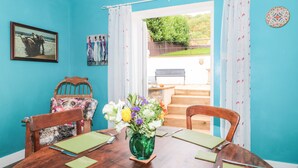 Comedor con mesa de madera dispuesta para cuatro personas, un jarrón de flores y vista a un jardín a través de un acceso.