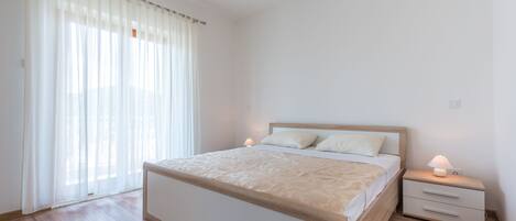 A bedroom with a wooden bed, white curtains, a bedside table with a lamp, and a wooden floor.