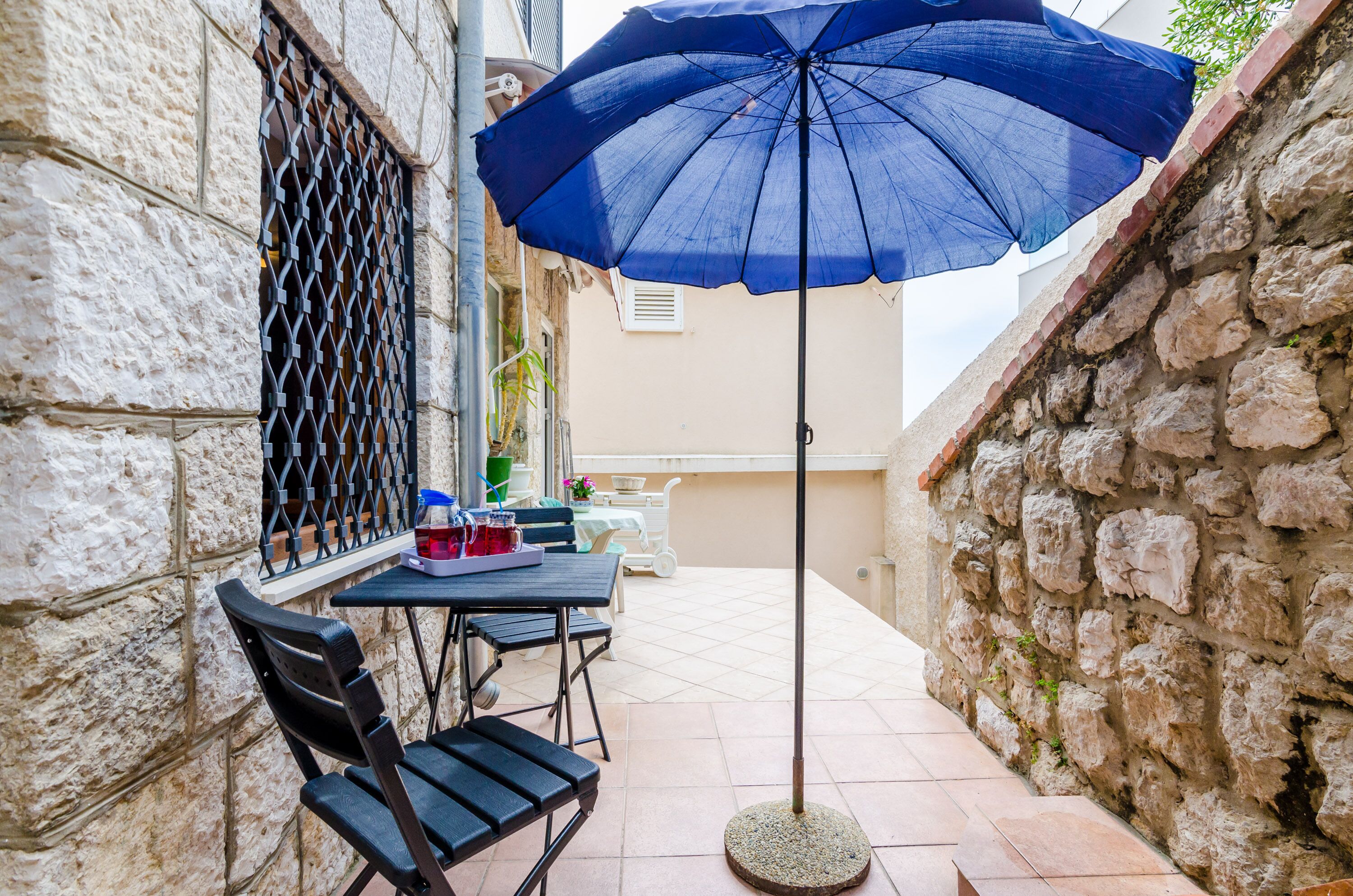 Un pequeño patio al aire libre con una mesa, sillas, un paraguas azul y un muro de piedra.