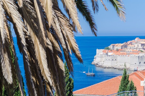 A coastal town with a historic fortification, a boat on the water, and palm trees framing the view. - Apartments Davor (Dubrovnik)