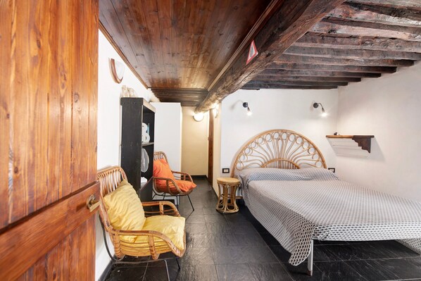 Una habitación con techo de madera, una cama con colcha estampada, un sillón de mimbre con cojines, una pequeña mesa redonda y un estante fijado a la pared.