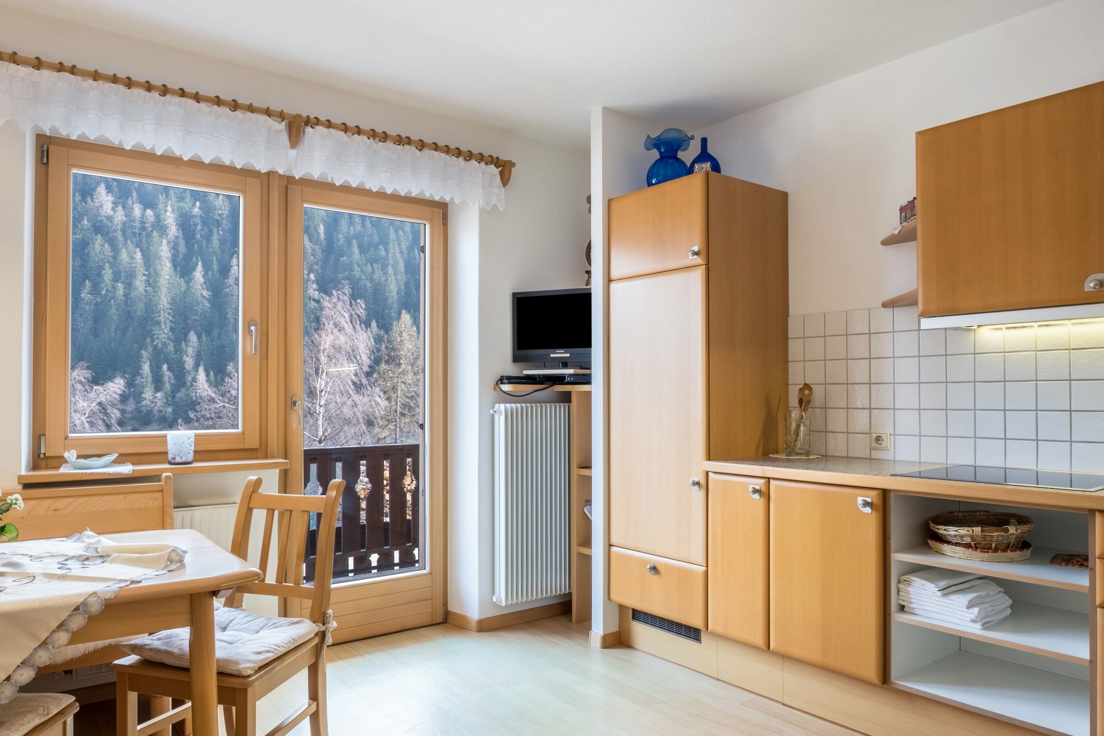 A kitchen with wooden cabinets, a dining table with chairs, a TV, and a view of trees through the window.