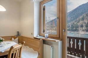 A room with a wooden table, chairs, a window with a view of a forest and mountains, and a balcony with a railing.