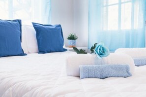 Un lit bien fait, avec une literie bleue et blanche, une petite plante en pot et une rose bleue posée sur les oreillers.
