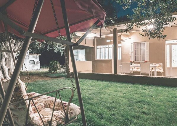 Une terrasse avec un fauteuil suspendu et une table entourée de chaises, un espace abrité, et un bâtiment avec des fenêtres et des portes en arrière-plan.