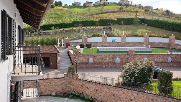 A residential building with a balcony, brick walls, a green lawn, and a hillside with houses.