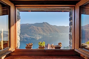 Une fenêtre donnant sur un lac et des montagnes, avec un cadre en bois et des plantes en pot posées sur le rebord.