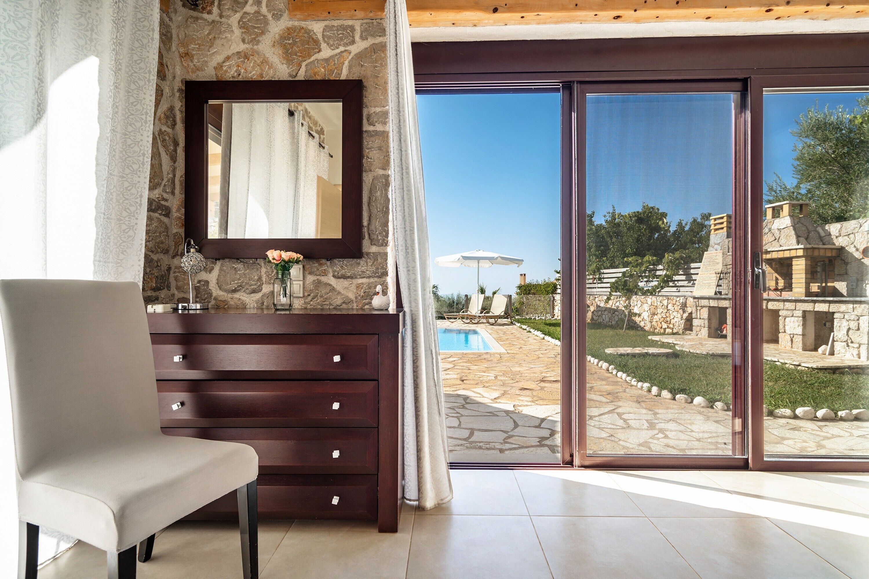 A room with a stone wall, a wooden dresser, a chair, a large sliding glass door leading to a pool area.