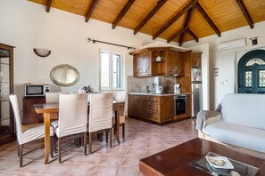 A living room with a sofa, a dining table with chairs, a kitchen area with wooden cabinets, and a tiled floor.