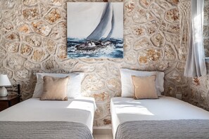A bedroom with two beds, a stone wall, a painting of a sailboat, and a nightstand with a lamp.